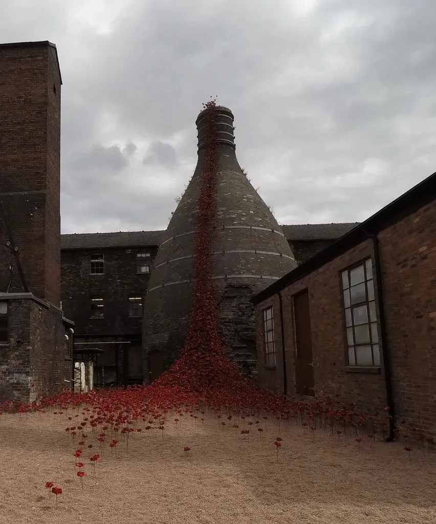 A large, cylindrical brick structure resembling a kiln or chimney stands prominently in the center of an industrial courtyard. The structure is adorned with a cascade of red poppies that flow down its side and spill onto the ground, creating a striking contrast against the muted tones of the surrounding brick buildings. The scene is set under a cloudy sky, giving it a somber yet poignant atmosphere. The red poppies symbolize remembrance and are scattered across the sandy ground, extending outward from the base of the central structure. The overall composition blends industrial architecture with a powerful artistic installation.