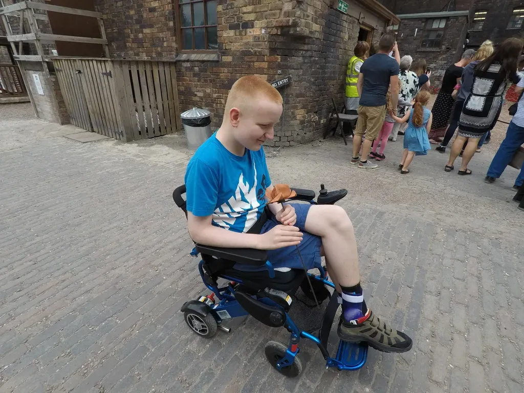 George, a young boy with short, light hair, sits in a blue and black motorized wheelchair on a brick-paved street. He wears a blue t-shirt with a white design, blue shorts, and brown shoes, and holds an orange poppy ornament in his hands. In the background, a crowd of people, including adults and children, are gathered near a brick building, observing the poppy field.