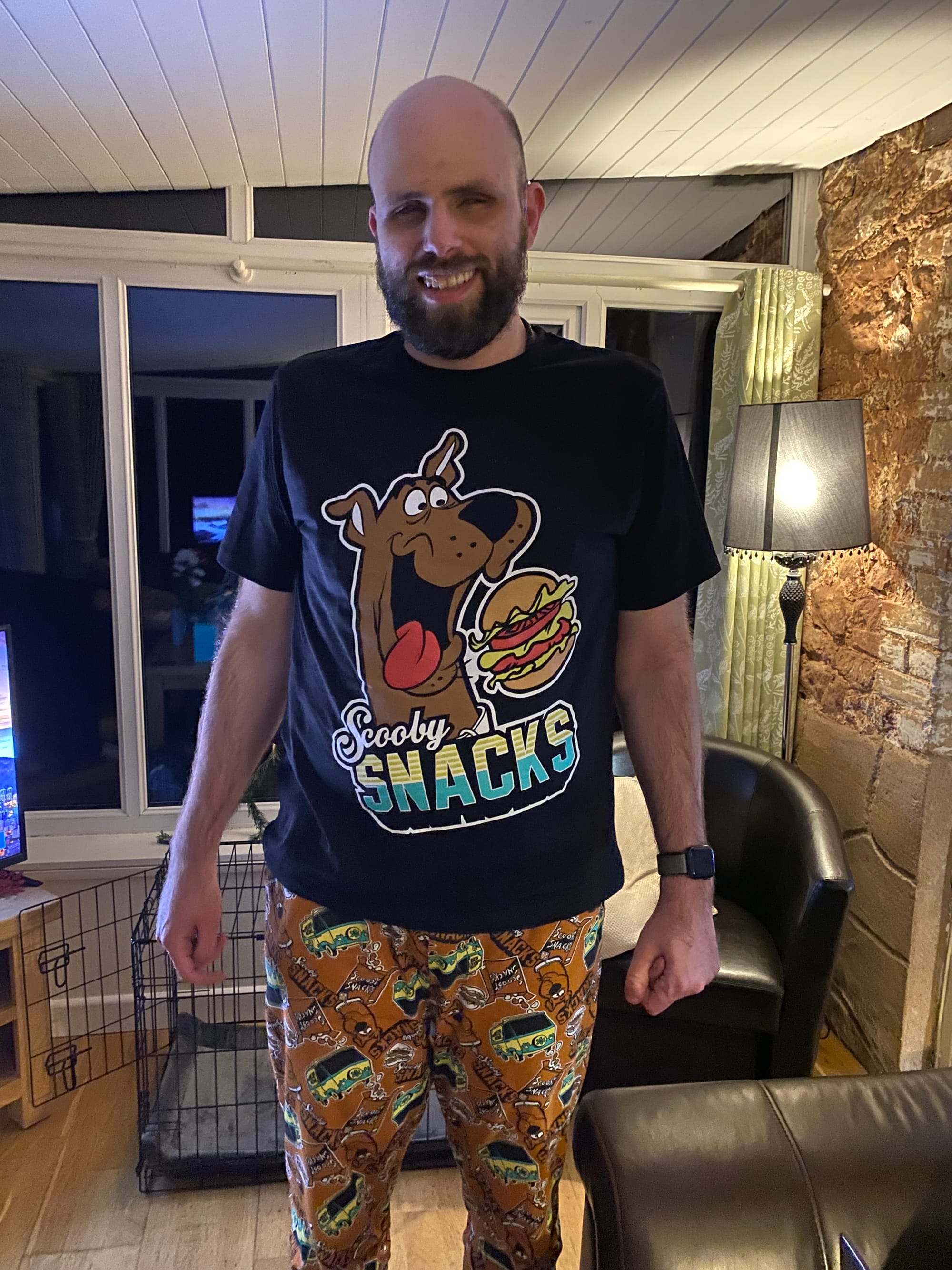 A bearded man standing in a living room. He is wearing pyjama trousers that have images of Scooby Doo all over them, and a pyjama top that has a picture of Scooby Doo with a hamburger. It has the words Scooby Snacks.