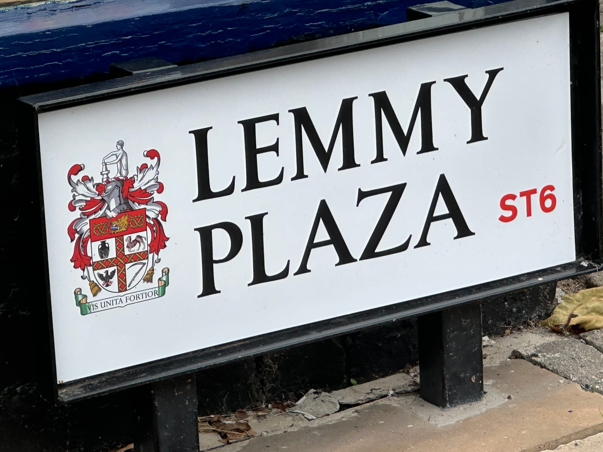 The image shows a street sign. On the left is the coat of arms for the City of Stoke-on-Trent. On the bottom right there is text, printed in red, that says ST6. In the middle, in black capital letters, is text that says Lemmy Plaza.