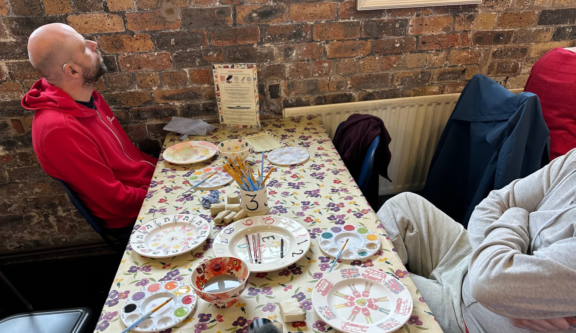John and George are sitting at a table with a floral cover on it. The three painted plates are visible, along with three paint palettes, two water bowls, a pot with the number 3 painted on it, containing paintbrushes, some sponges and a plate showing the colours the glazes would be when fired