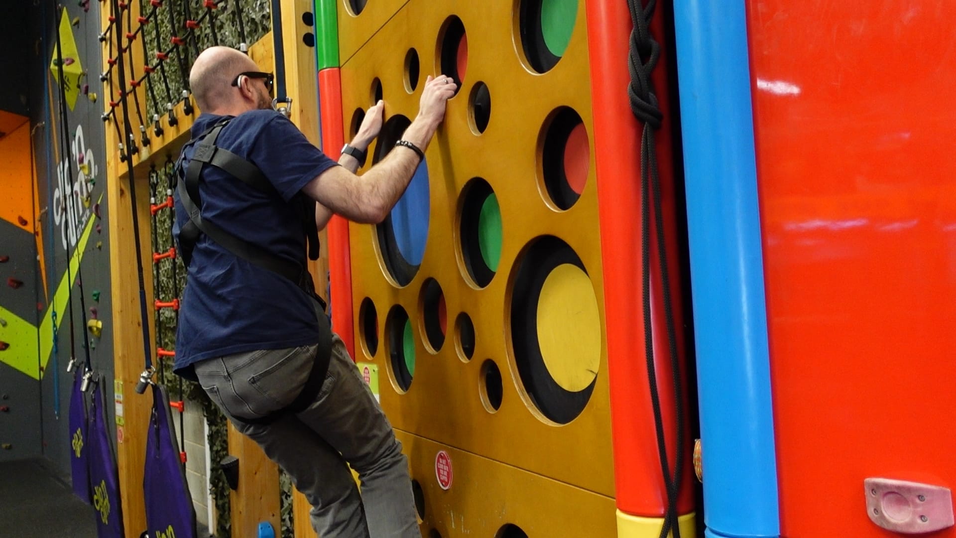 John climbing a yellow wall, with different size holes in it for hand and foot holds. The insides of the holes have different colours. John is wearing his Meta AI glasses so he could get some point of view footage