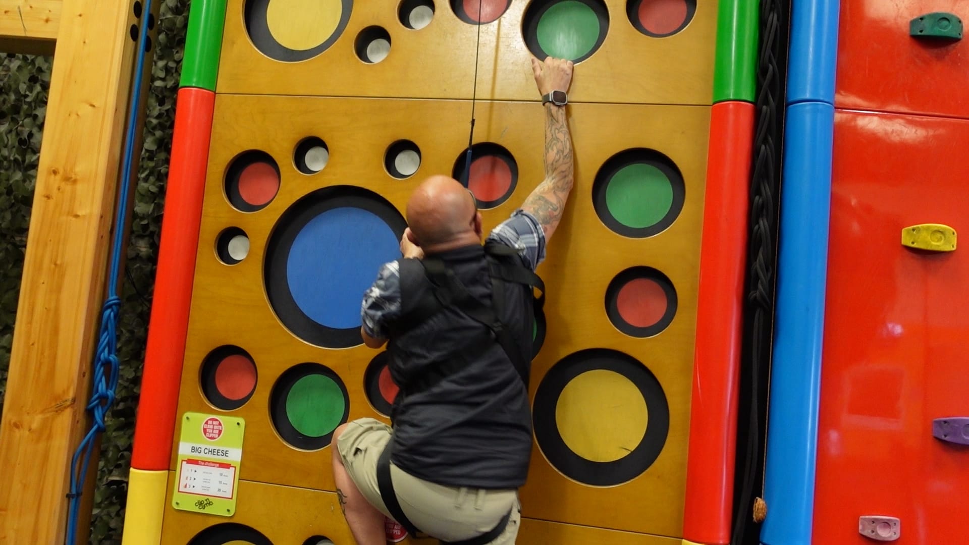 Wayne climbing a yellow wall, with different size holes in it for hand and foot holds. The insides of the holes have different colours. 