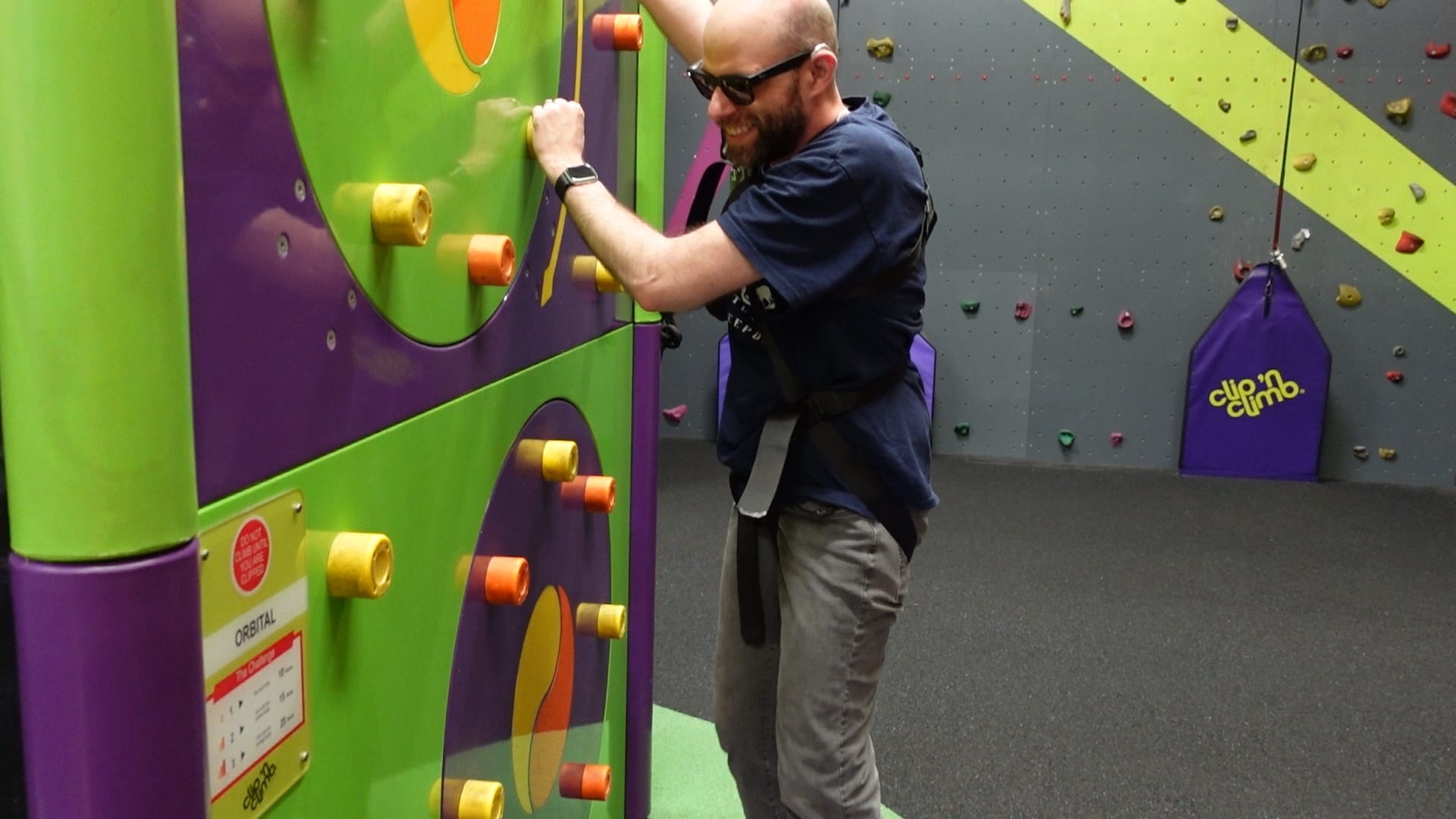 John is standing in front of a wall with hand and foot holds arranged in a circle. His left hand is on a hand hold and his right hand is out of shot. The circle these hand holds are in spins when weight is applied. 