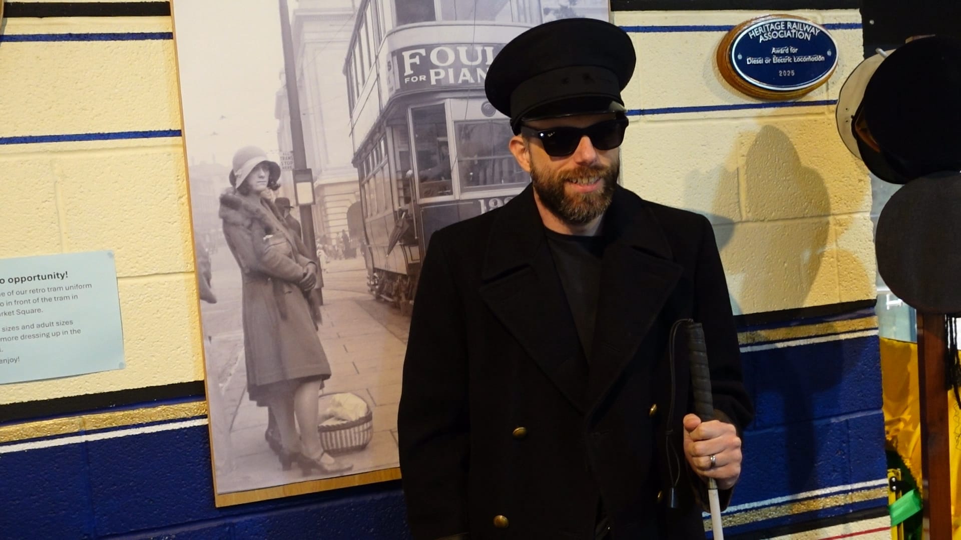 John wearing a dark peaked cap and jacket. He is wearing dark glasses and holding his cane in his left hand