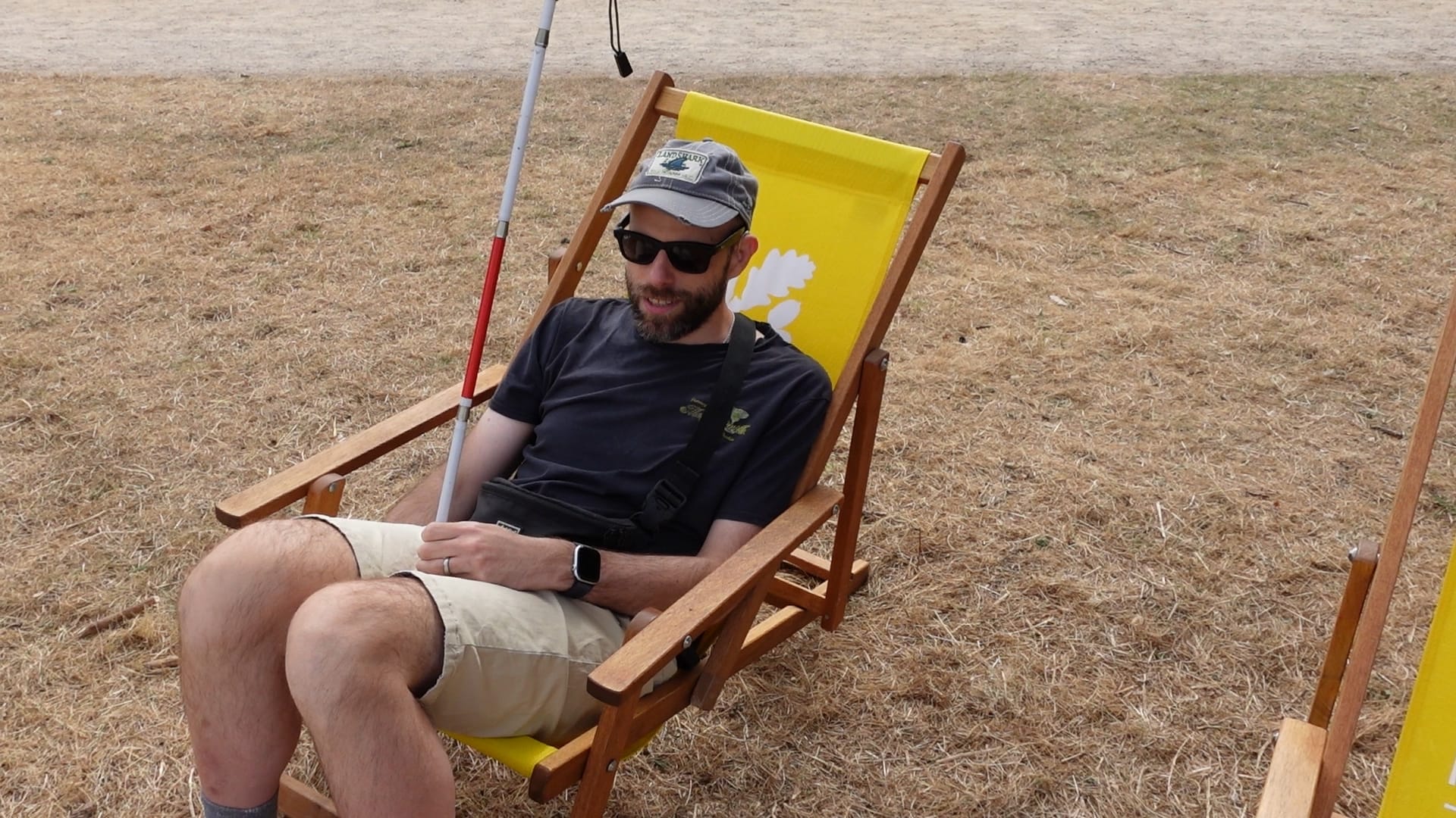 John sitting on a bright yellow National Trust deckchair. He is holding his unfolded long cane in his left hand. The deckchair is sited on some scorched grass