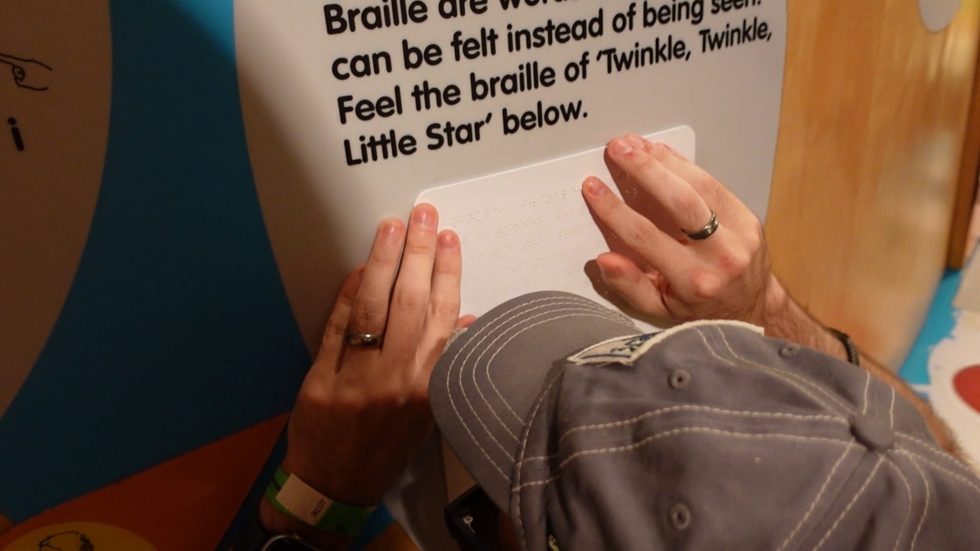 John is crouched down, feeling the Braille on the wall in front of him. Above the Braille we can see the printed words "Feel the braille of 'Twinkle, Twinkle, Little Star' below."