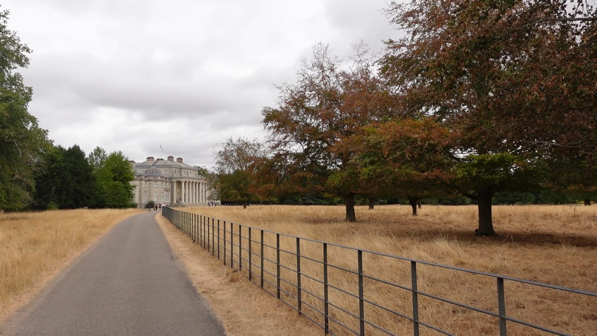 Approaching the mansion from the right along a long path. There is scorched grass on either side, and a fence on the right of the path. There are trees on either side too, providing the only bit of greenery