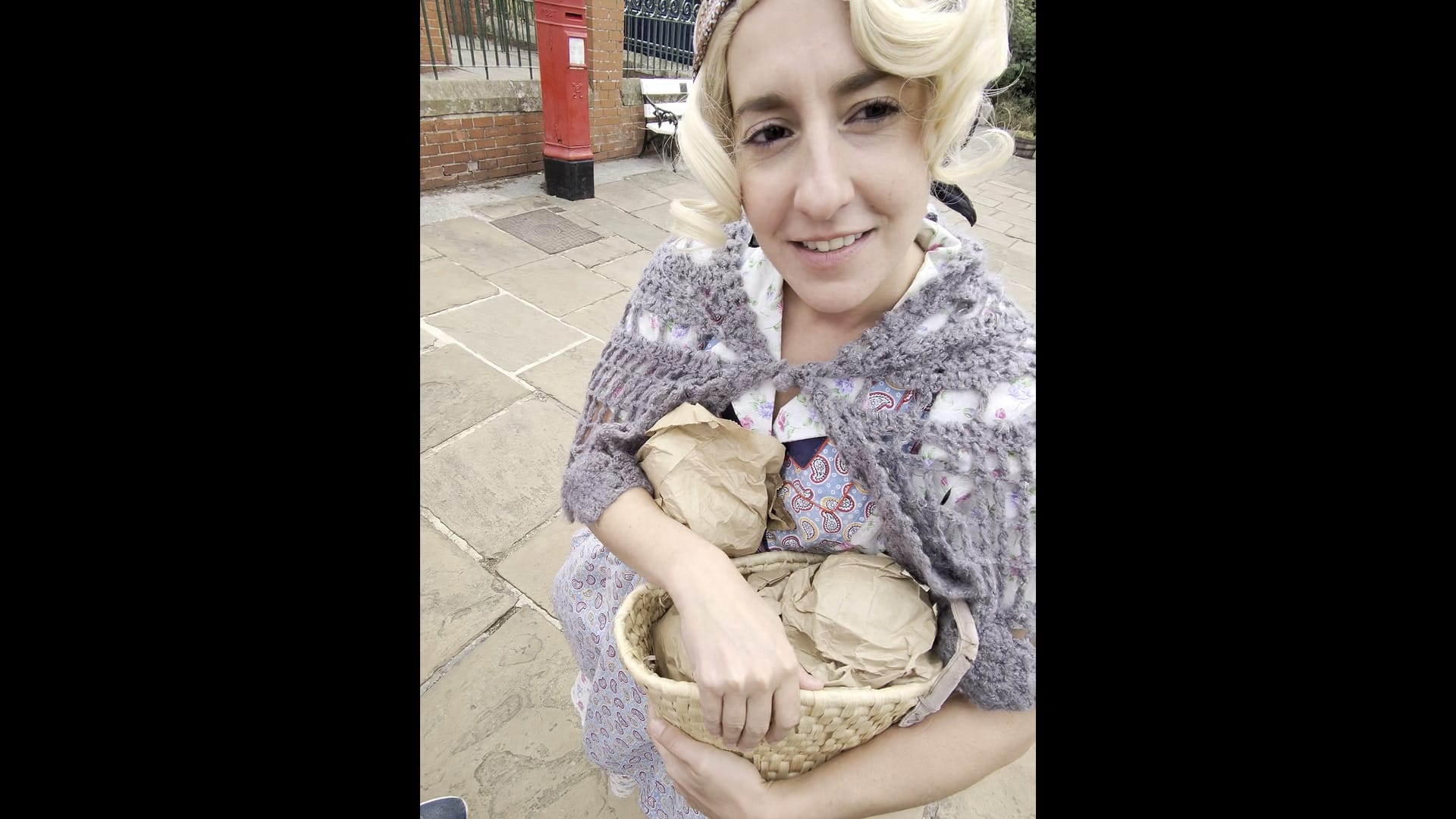 A blonde haired woman dressed in 1936 period costume, carrying a basket of items in brown paper bags