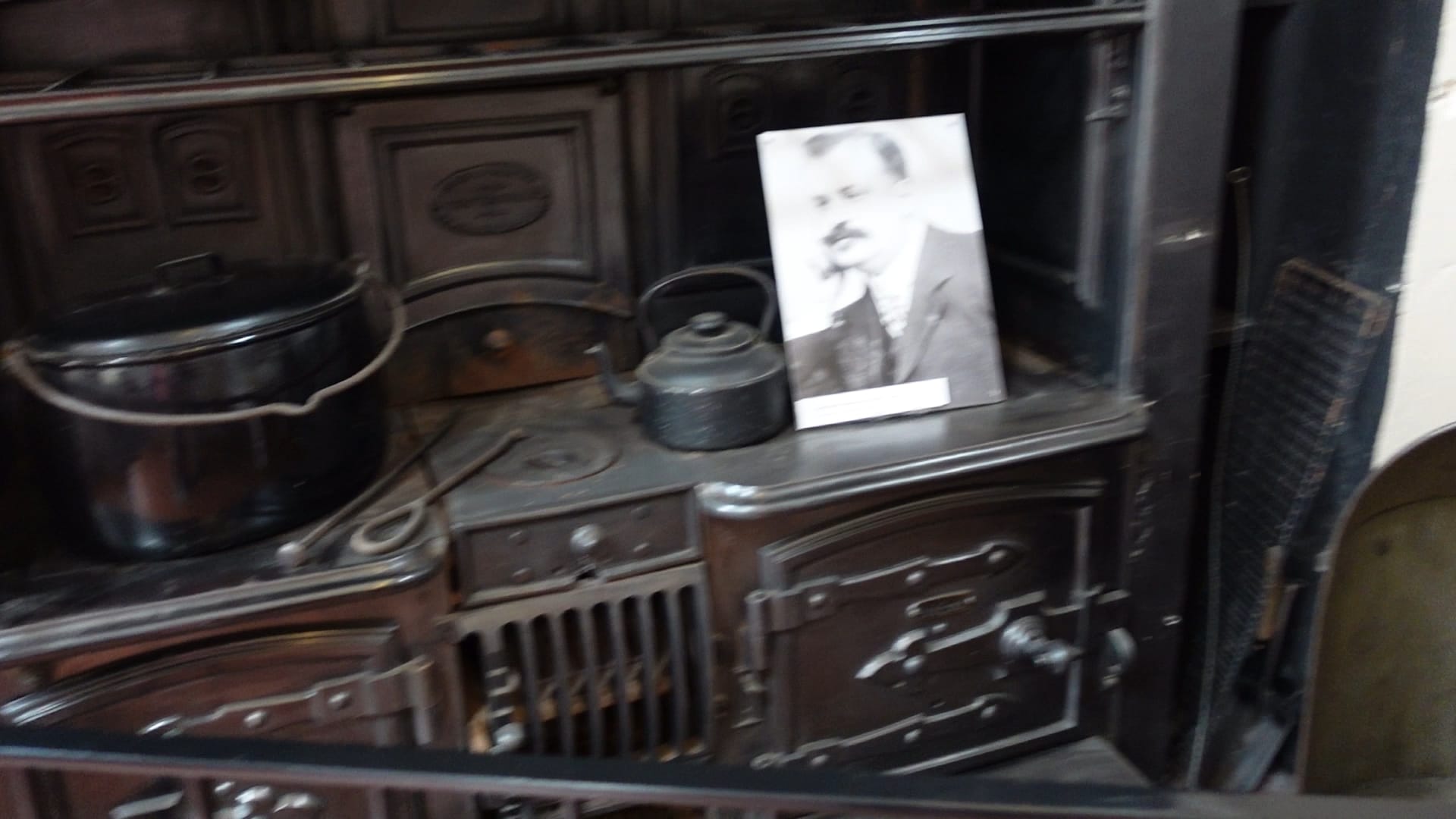 A large black range cooker. There is a pot on it, on the left. Towards the right there is a kettle and on the right is a photograph of a man - possibly the chef