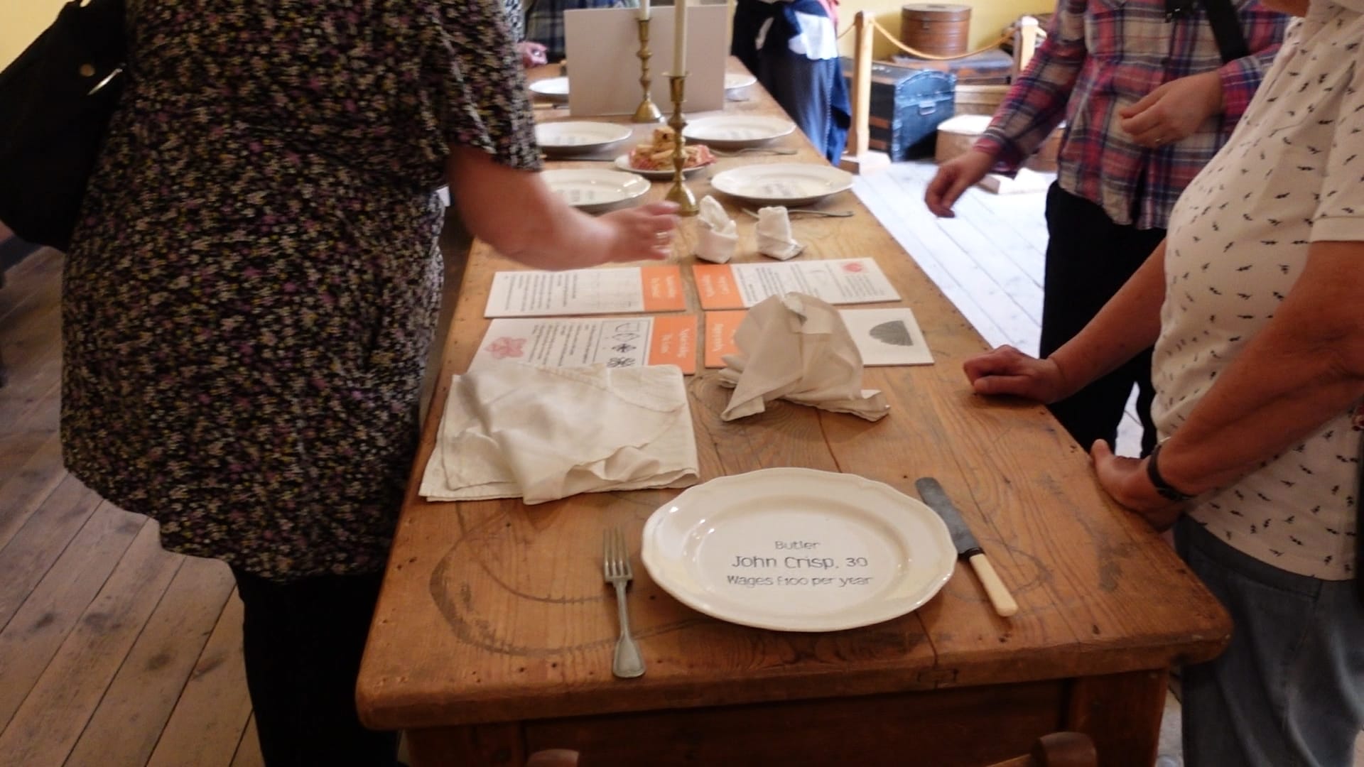 A long wooden table with plates on denoting where each servant sat, their name, their age and their pay for the year. The plate we can see at the head of the table says "Butler, John Crisp, 30, wages £100 per year"