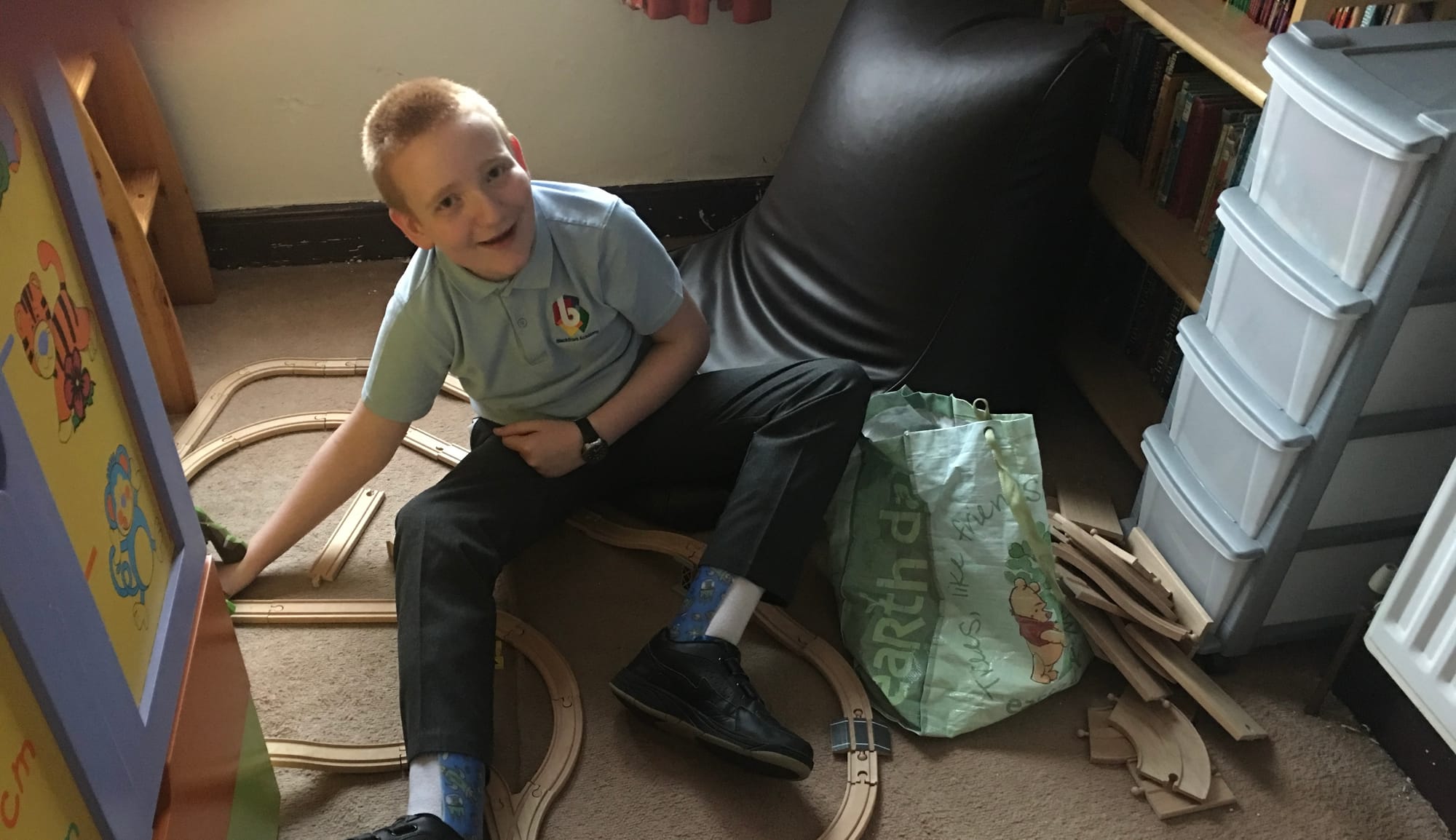 George, aged around 11, sitting on the floor playing with his wooden train set. He is smiling at the camera, and his blue splints can be seen on his legs