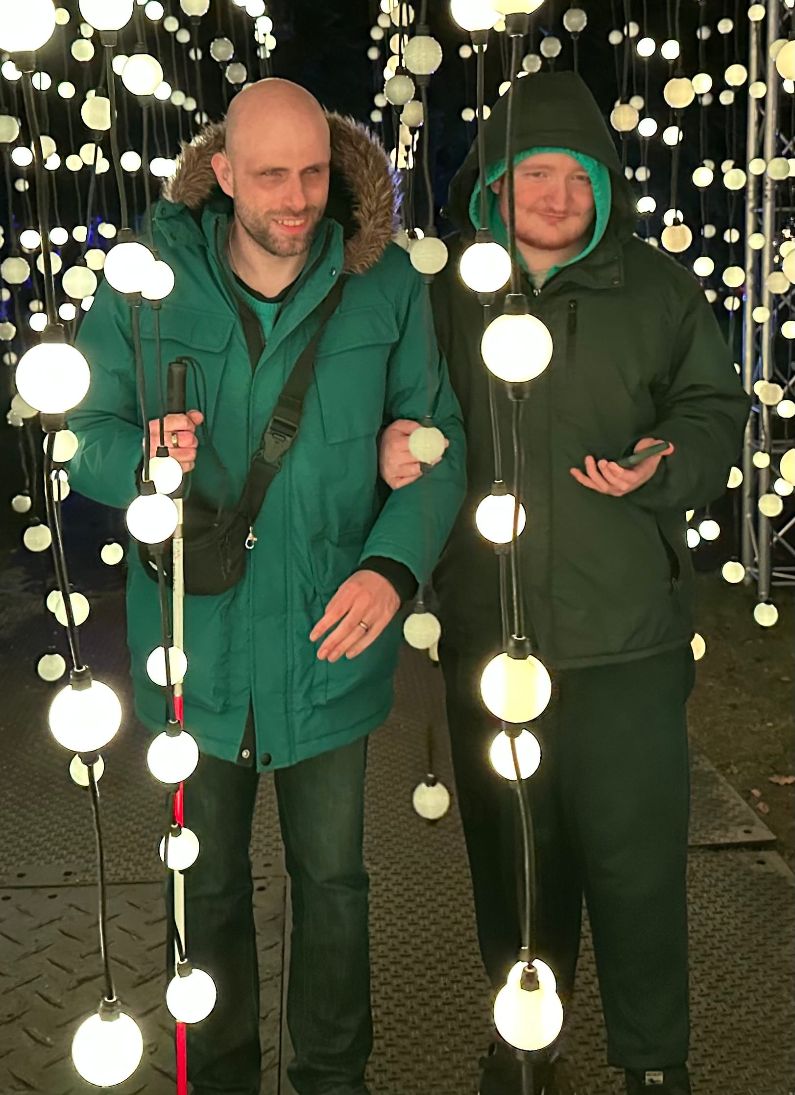 George is standing on John's left, holding his arm. They have lights all around them and hanging down in front of them