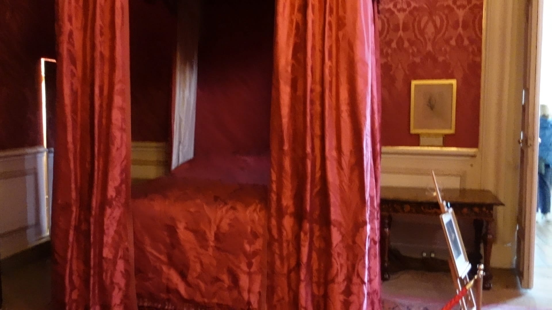 A four poster bed with red covers and red curtains around it. The wallpaper behind it is also red.