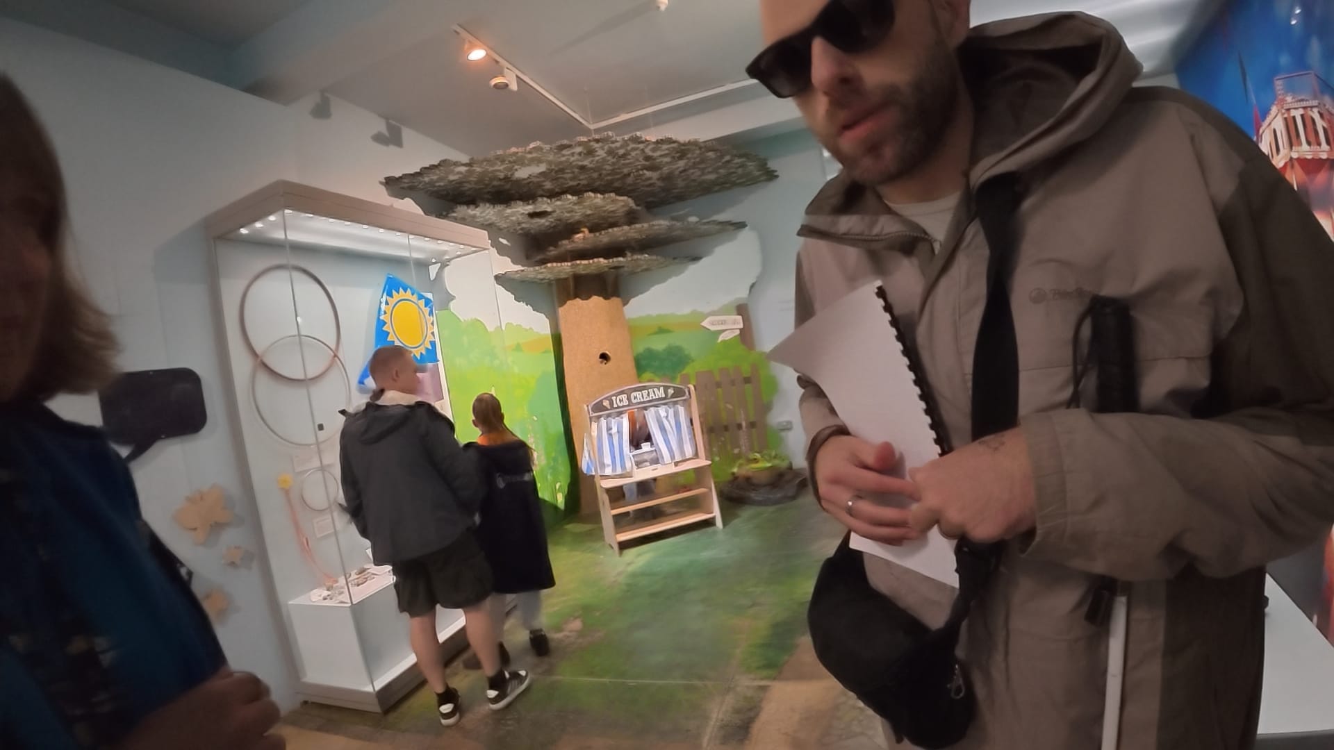 John is wearing his dark Meta glasses and a raincoat. He has a bag across his shoulder and is holding his long cane under his left arm. He has the Braille guide against his chest. His right hand is reading the dots and his left hand keeping his place.
