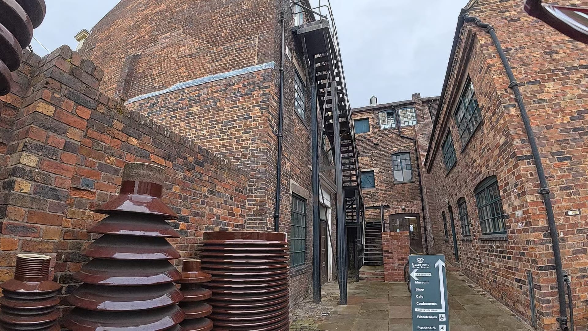 A collection of artefacts from the museum are on the left. In front there are buildings and a sign pointing out where the main entrance is, and the ramped entrance via the shop