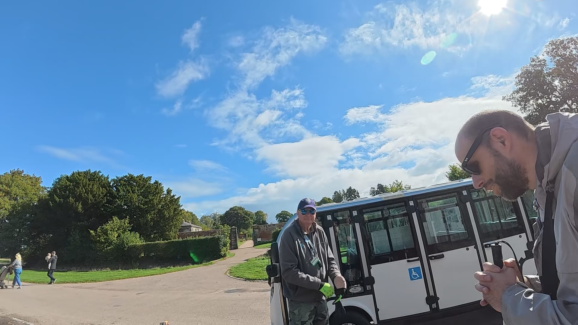 A picture of the accessible vehicle, Ernie, with the driver standing beside it. John is standing to the right of the picture wearing his Meta glasses and holding his cane.