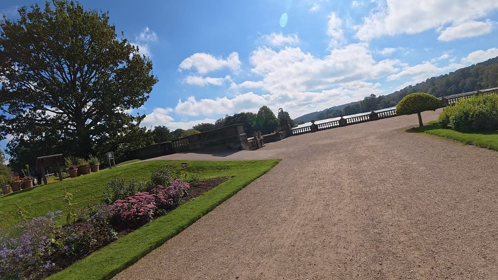 A view along a path towards the lake. We can see trees in the distance and some flower beds to the side of us.