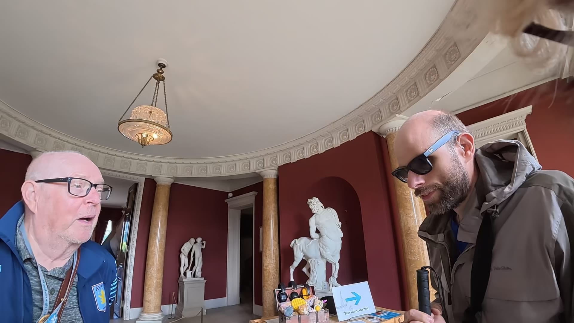 Just inside the entrance hall. There are white statues in the background. On the left is a volunteer who checked our membership cards and told us a little bit of the history the mansion. John is standing on the right, wearing his Meta glasses and holding his cane, listening to what is being said.