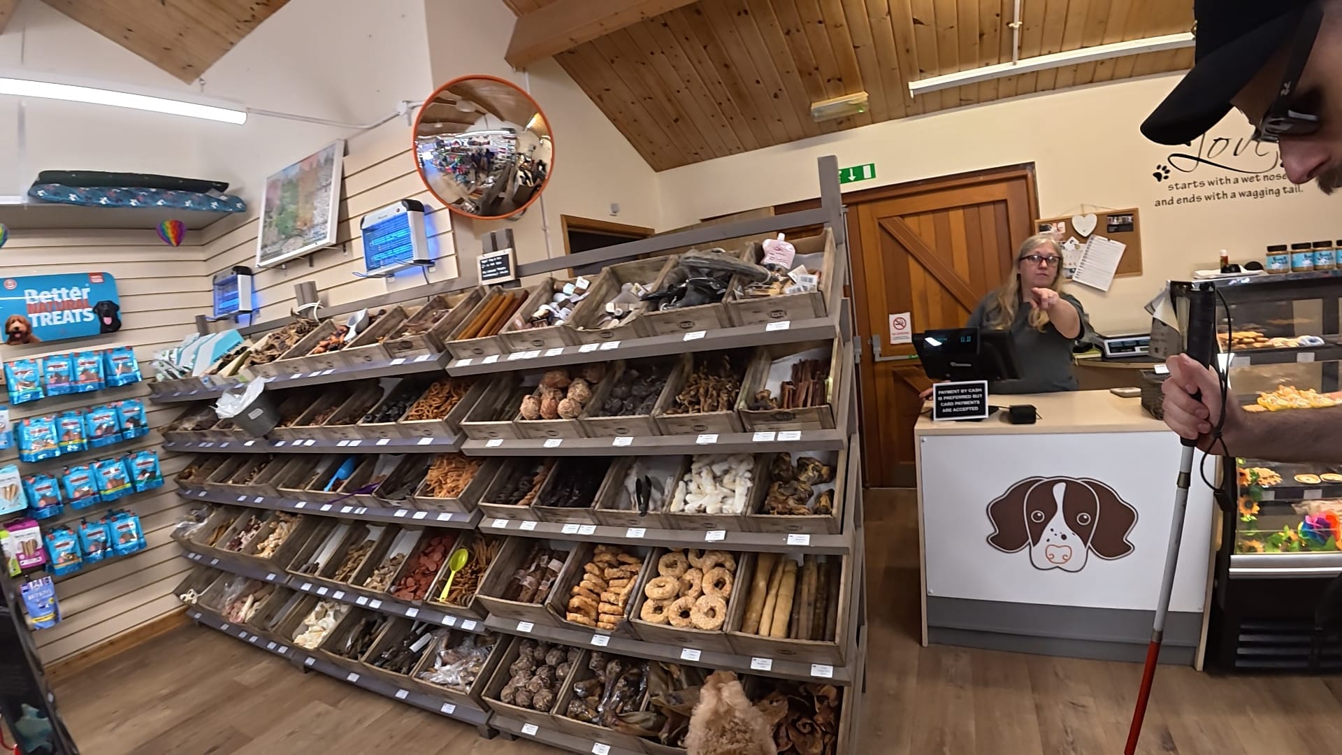Near the counter of the dog shop. a woman behind the counter is pointing out where the poo bags are. Meanwhile, Jasper is by a display of dog treats, checking them out