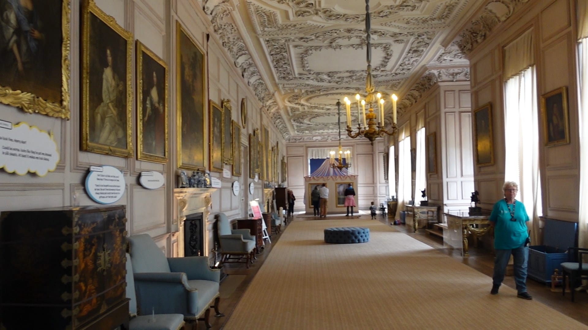 The long gallery has paintings down the left side and windows on the right. The ceiling has ornate carvings
