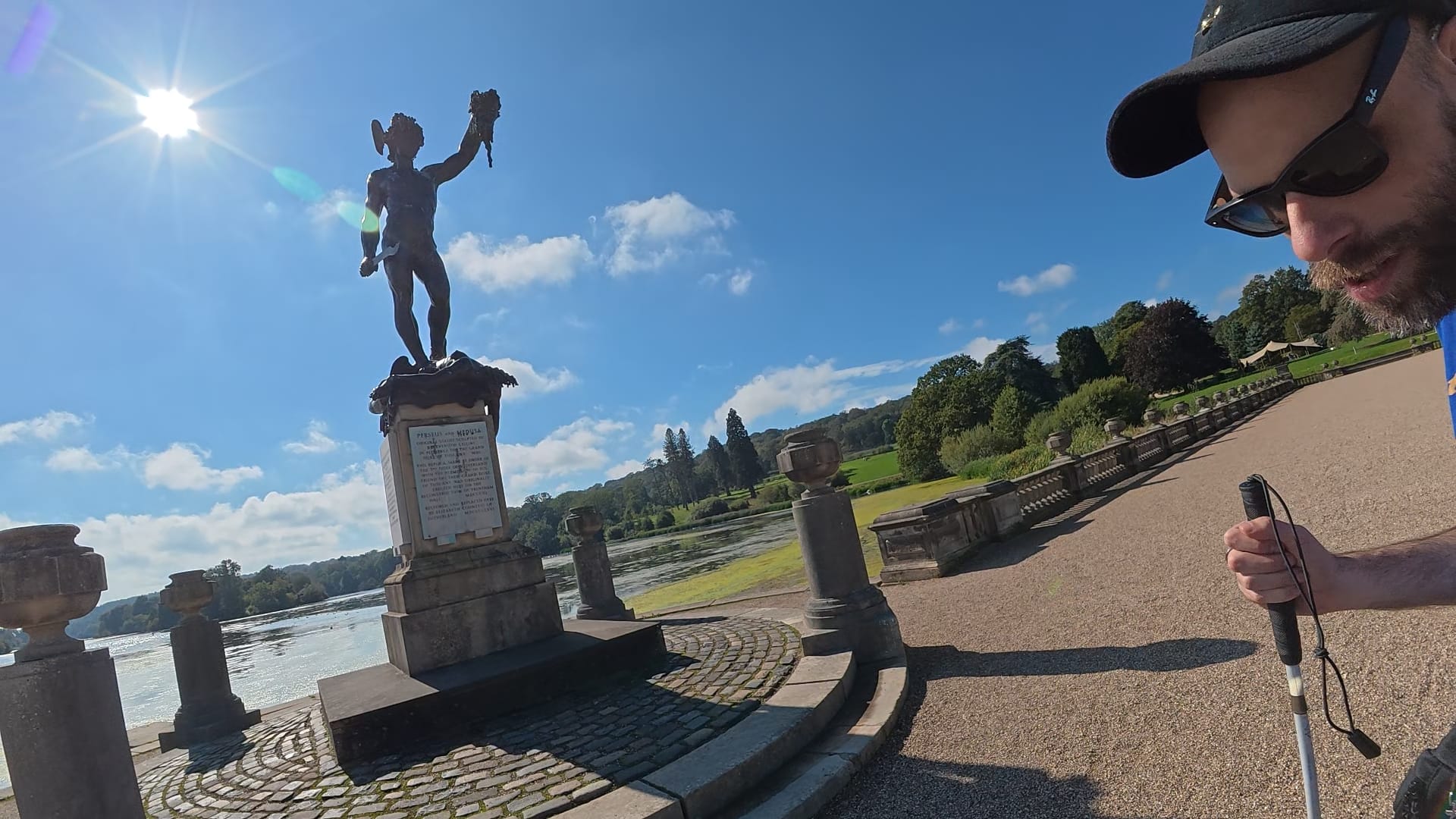 The Perseus and Medusa statue standing in front of the lake. John is standing to the right the picture, with his white cane in his right hand. He is looking slightly towards the camera as he listens to the inscription being read.