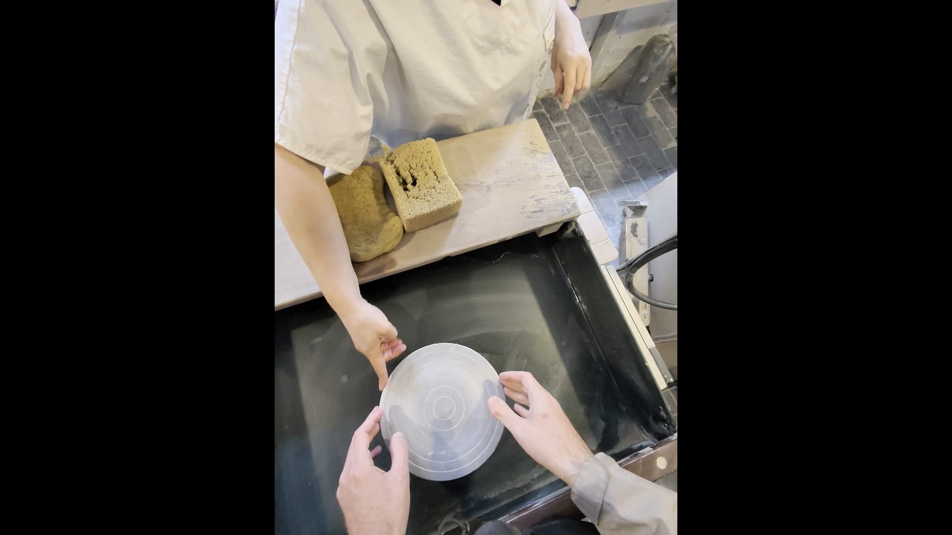 We can see John's hands touching a small potter's wheel. A staff member is in front of him pointing out different parts of the wheel. There are some sponges in front of her.