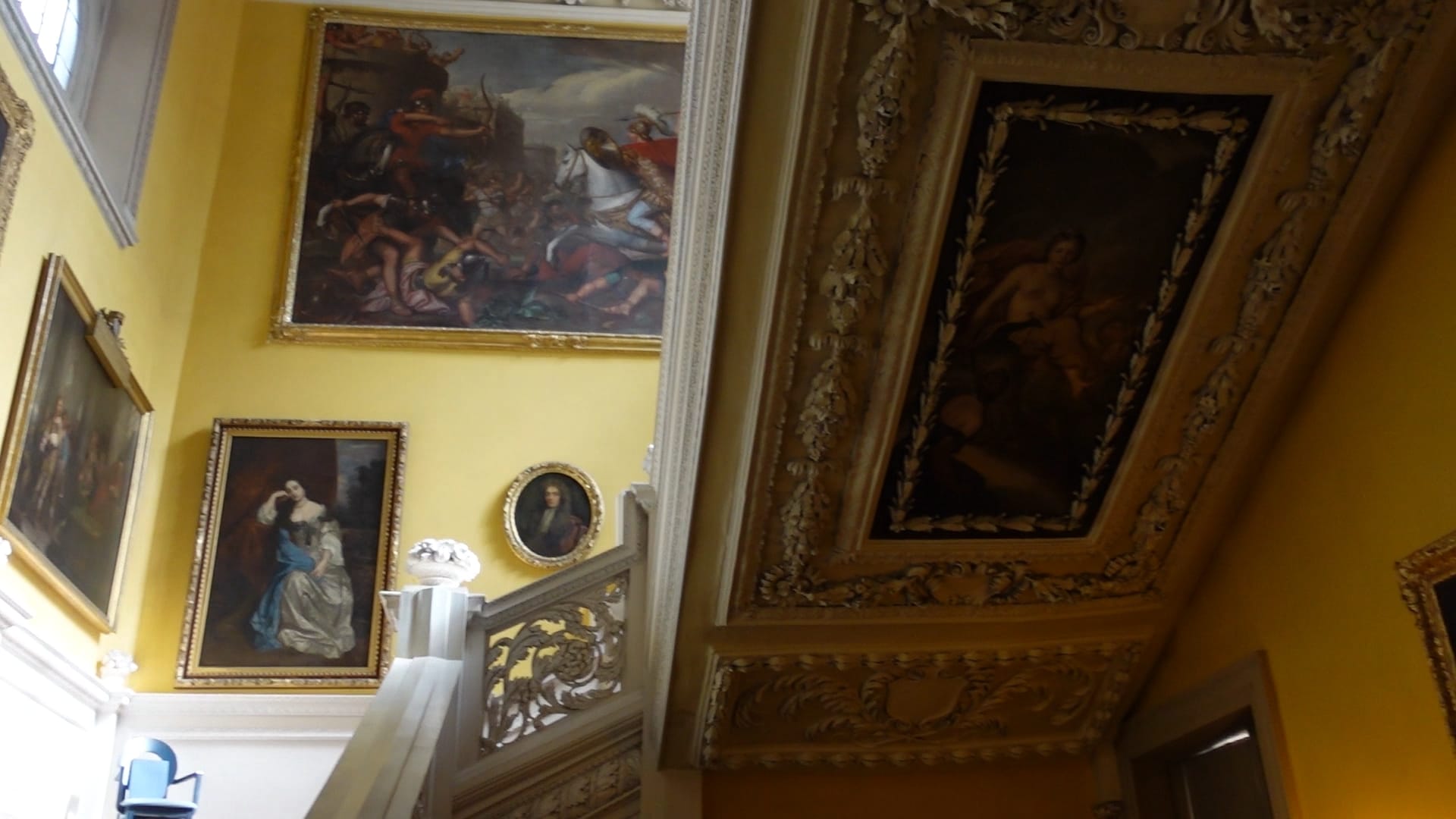 A grand, white staircase, with paintings going along the side. Underneath the stairs is another painting, with ornate carving around it.