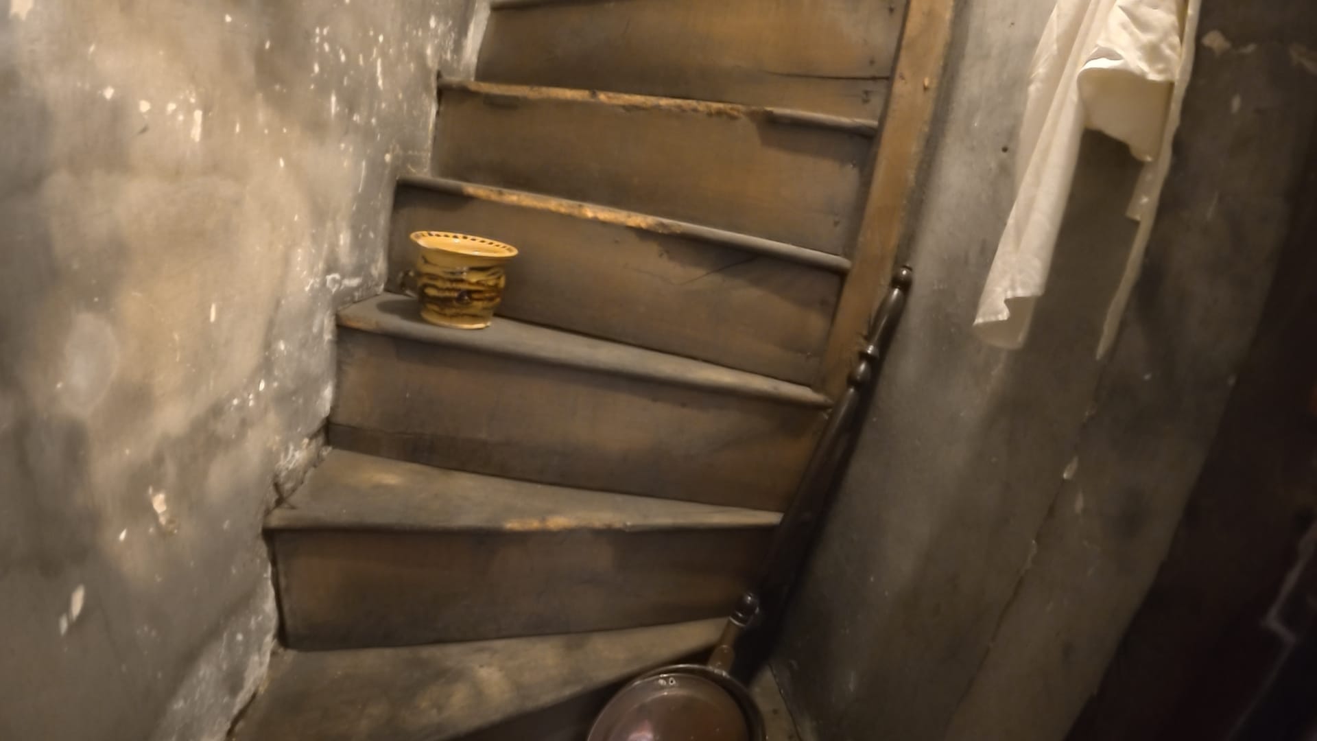 An old wooden staircase hidden behind a door. It is steep and twisty, with narrow treads. A bed pan is leaning against the front of the stairs and a pot is on the third stair up
