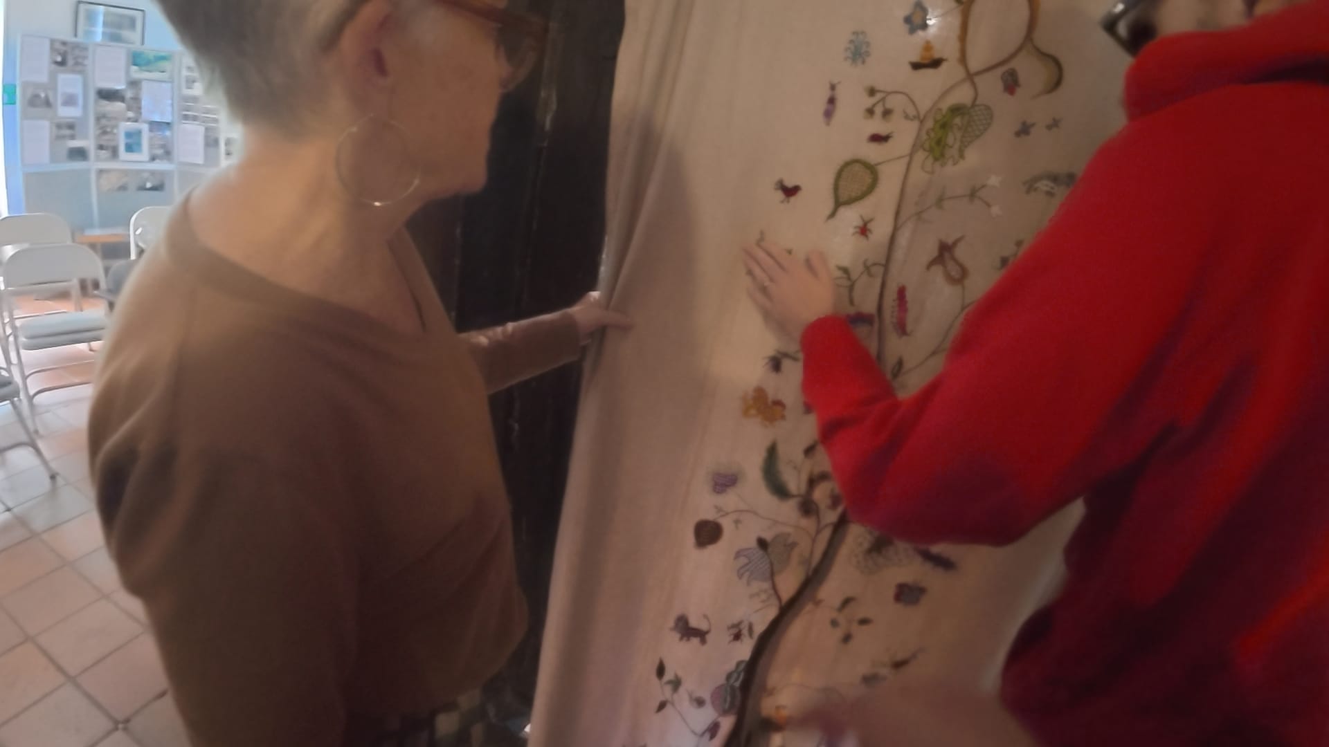 A large embroidered piece of the Tree of Life hangs in a doorway. A volunteer stands to the left, watching John on the right feeling the embroidery