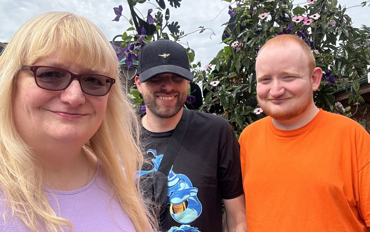 Three people in a photo. A woman with long blonde hair is on the left (Sue), a man wearing a black cap and shirt is in the middle (John), and a man in an orange shirt is on the right (George)