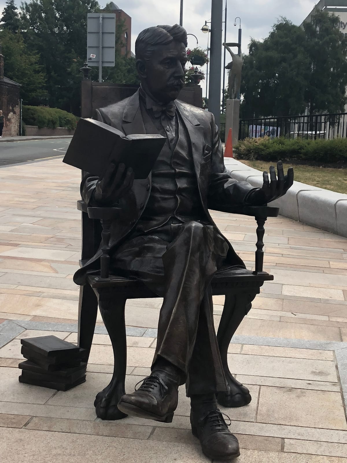 A statue of Arnold Bennett, seated in a chair, holding a book in his right hand. His left leg is crossed over his right and he has a pile of books on the floor to his right