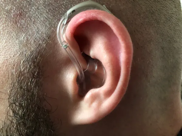 A close-up of a John's ear wearing a behind-the-ear hearing aid, with the device resting on the ear's outer curve and a clear tube connecting to the ear canal.