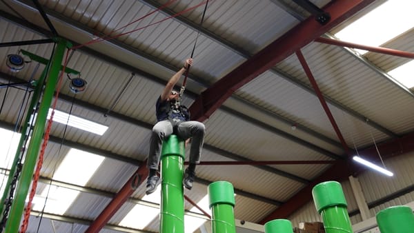 John is sitting on top of a green pole, holding onto a rope in front of him