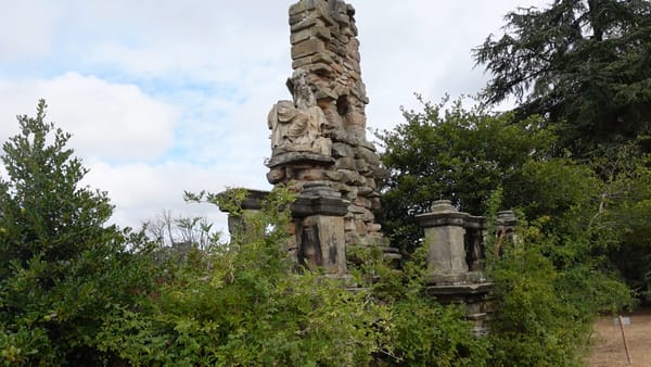 A tall, weathered stone monument with carved figures, part of a garden ruin at Shugborough Estate, surrounded by trees and shrubs.