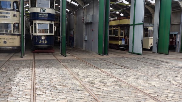 A tram shed, with different old trams inside it. There are cobbles and tram lines in front of the shed