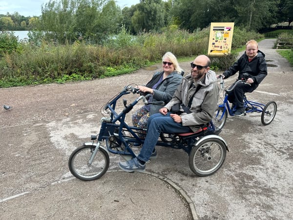 John and Sue are sitting on a side by side tandem. George is behind on a trike. Westport Lake is in the background