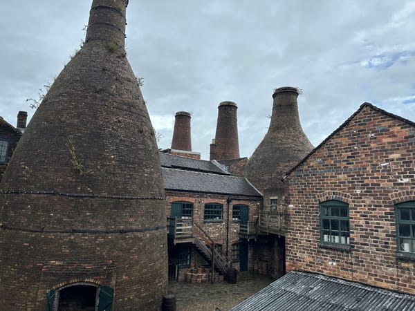 In front, slightly to the left is a bottle oven. Across the courtyard is another. There are buildings in between and other chimneys in view.