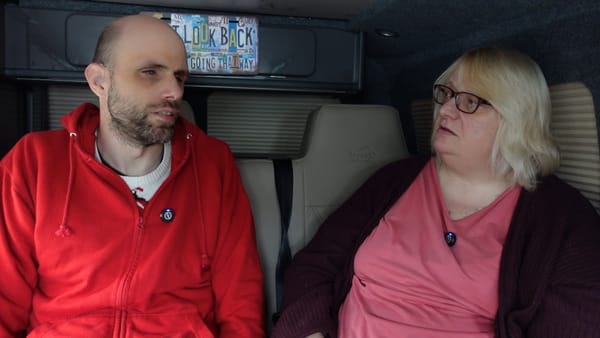 John on the left and Sue on the right, in the camper van. John is talking and Sue listening