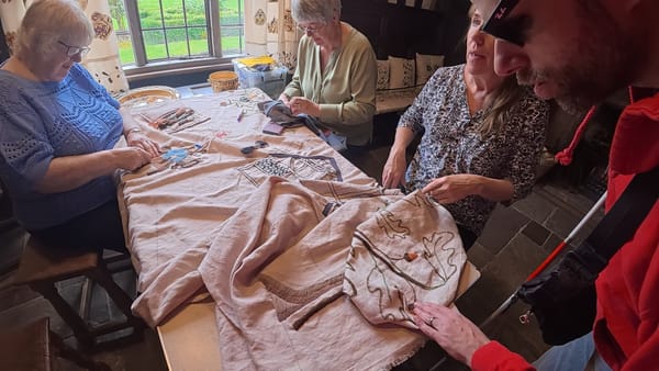 A large piece of embroidery work on a table. Three volunteers are seated, while John feels their work